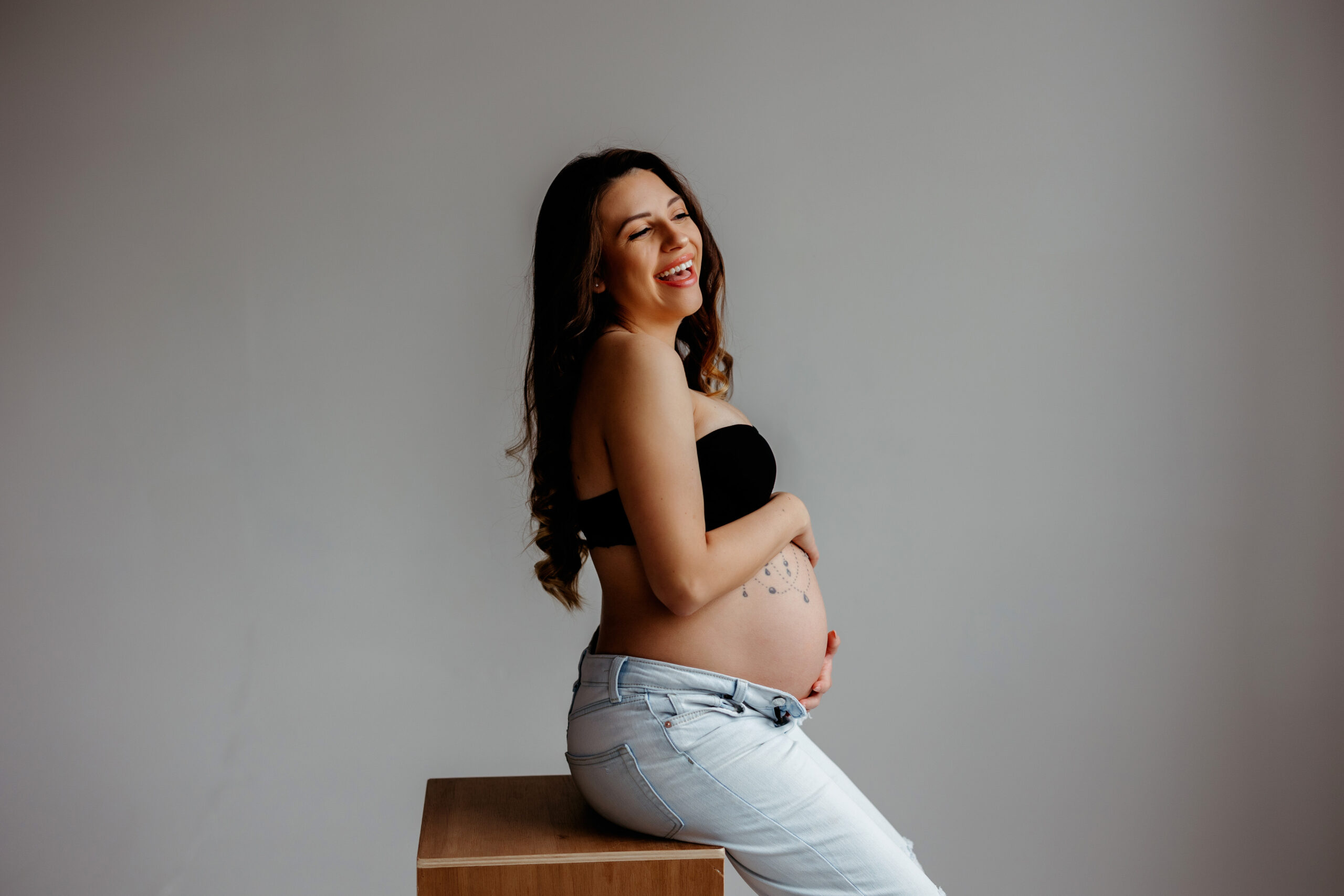 Pregnant mom shows off baby bump wearing a black bralette and jeans for her indoor maternity photos in Denver